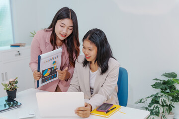 Two Asian businesswomen working together at a desk, focused and professional. They collaborate on...
