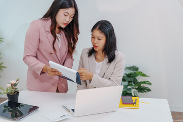 Two Asian businesswomen working together at a desk, focused and professional. They collaborate on...