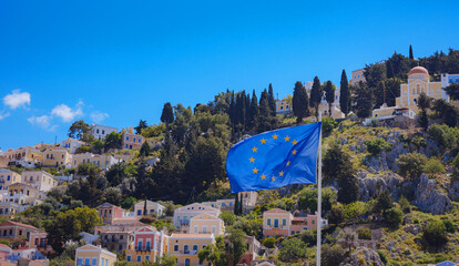 View of EU flag waving in air serene Mediterranean vibe.EU flag European Flag It features a circle of 12 gold stars on a blue background. in harbor town of Symi (Symi Island, Greece)