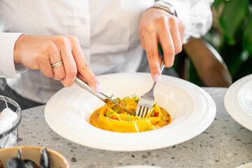 A close-up of hands using a fork and knife to cut a plate of colorful pasta, showcasing its vibrant ingredients and elegant presentation in a stylish dining environment.