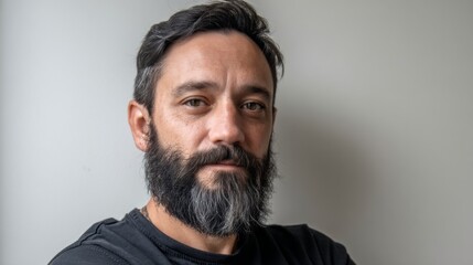 Fototapeta premium Standing confidently, a man with a neatly trimmed beard dons a black shirt. He exudes a sense of contemplation, framed by a simple yet modern background that highlights his features