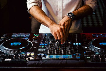Close-up of male DJ mixing music on a professional sound console in a dimly lit club setting,...