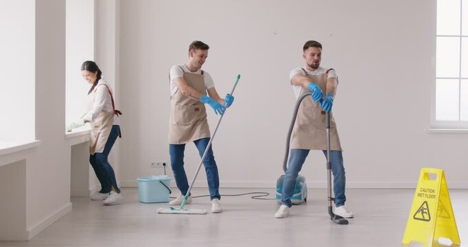 Work is pleasure. Cleaning company employees add little dance to their workday by performing cleaning tasks. Three cheerful cleaners in aprons and gloves are having fun while cleaning an empty room.