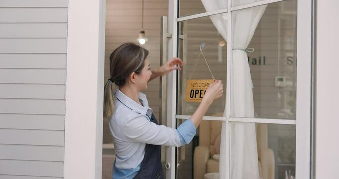 Asian adult woman with ponytail wearing apron standing outside spa salon smiling happily while hanging welcome open sign on glass door, sunny day background creating positive energy for business.