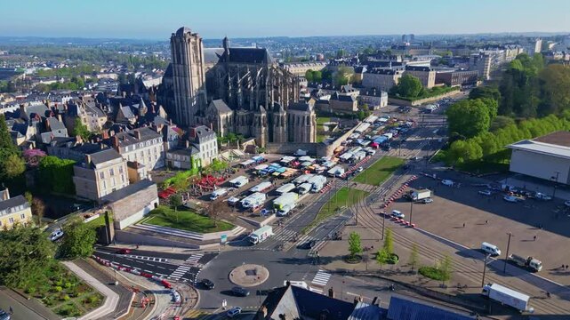 Forward drone motion over roundabout and Jacobins Market, leading to Le Mans Cathedral of Saint Julian under blue skies. France
