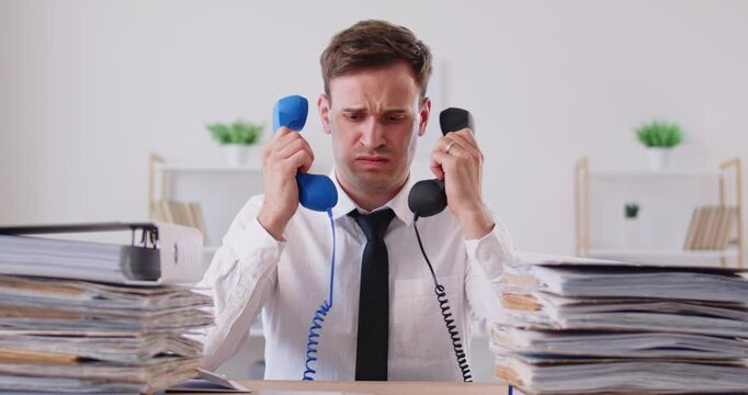 Stressed frustrated male company employee sitting at desk with two handsets in his hands, suffering from multiple calls. Overworked man amazed by stupidity of interlocutors, making displeased face.