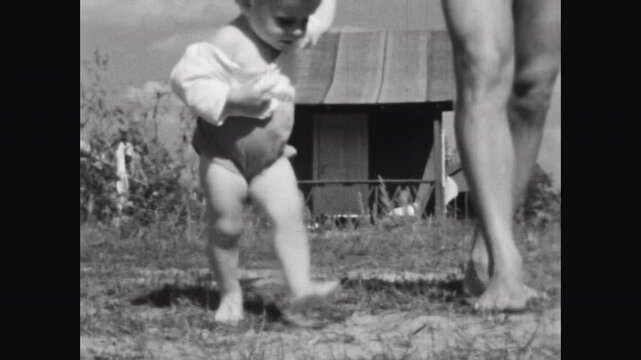 Close up, little child making first steps holding father hand on nature background. Cute toddler baby walk barefoot on grass. Vintage black white family film. Old archive video. Archival retro 1960s 