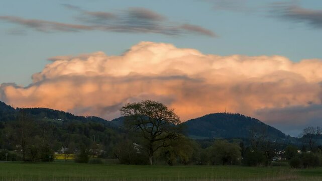 Clouds swirl and drift across the natural landscape at golden hour. The clouds are illuminated by yellow and pink. It's spring, and everything is blooming. Mountains can be seen in the background.