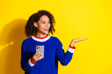 Young cheerful woman with curly hair holding a smartphone and gesturing with her hand against a...