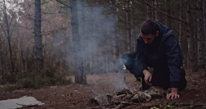 A man builds a campfire in the woods during winter. He carefully arranges kindling and uses a lighter to ignite the flames.