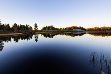 A peaceful sunset over a lake in Lapland, Sweden, with the reflection of trees in the still water.