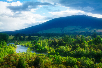 A vibrant landscape near Turangi, New Zealand, with a winding river flowing through dense green forest under the shadow of Mount Pihanga, bathed in dynamic light and dramatic cloud cover