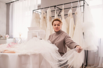 Obraz premium Portrait of seamstress in a wedding salon sews a wedding dress and veil, handmade, boutique. Small local business, self-employment.