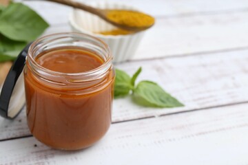 Tasty curry sauce in glass jar, powder and basil on white wooden table, closeup. Space for text