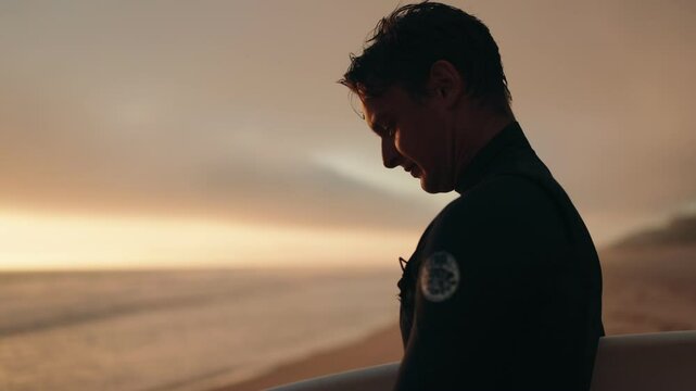 A surfer smiles as he prepares to surf at sunset. He is wearing a wetsuit and holding his surfboard on the beach, ready for the waves. The warm sunset colors create a serene and inspiring atmosphere.