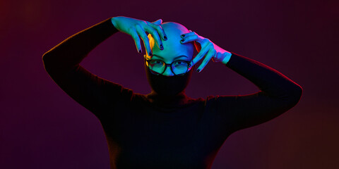Bald young woman in glasses raises arms framing face under vibrant lighting with expressive pose showing bold identity. Concept of self-expression, creativity, beauty, fashion, style, equality.