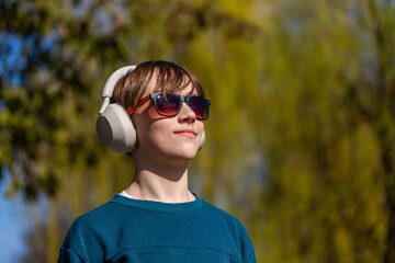 Portrait child little boy wearing sunglasses, headphones standing in a summer park and is smiling. Teenager boy with headphones listening to music on a mobile tablet, nature, outdoor, outside