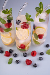 refreshing glass cocktails with lemon slices, raspberries, blueberries, and mint leaves in tall glasses on a light blue surface surrounded by scattered berries, capturing a vibrant and summery essence