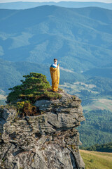 Young European man standing in a yellow sleeping bag on the edge of a rocky mountain peak,...