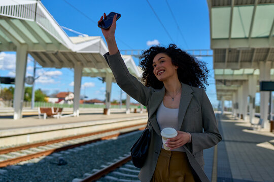 Young businesswoman with curly hair holding takeaway coffee and taking a selfie with smartphone while waiting for train at railway station on sunny day