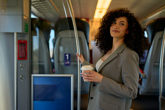 Businesswoman with curly hair holding disposable coffee cup and entering first class carriage of modern train, enjoying comfortable and convenient public transport for business travel