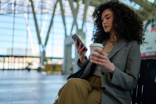 Young businesswoman sipping coffee while using her smartphone, patiently waiting for her flight in the airport terminal. Modern architecture and natural light create a stylish atmosphere