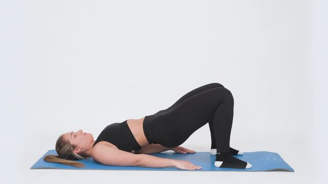 A focused woman in black sportswear engages in a glute bridge workout on a blue yoga mat on white background, demonstrating strength and form