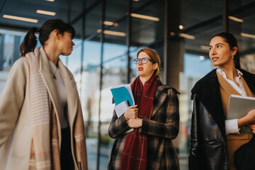 Three professional women engaged in conversation outside a modern glass building, symbolizing teamwork.