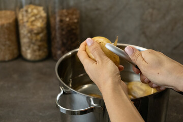 Closeup of hands peeling potato above steel pot in kitchen