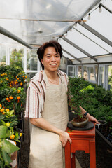 Cheerful asian florist holding bonsai tree on table in greenhouse