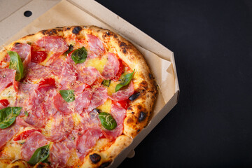 Pizza with cheese, ham, green basil leaves and tomatoes in paper box on black background. Food product advertising banner, copy space