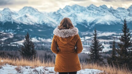 A woman in a thick winter coat stands atop a snowy hillside, gazing at the sweeping view of snow-capped mountains, feeling the peaceful solitude of nature.