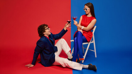 Happy man and woman raising glasses of cocktails in toast, seated against split red and blue studio background. Concept of human emotions, relationship, lifestyle, party, celebration