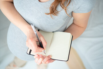 Future mother writing detailed maternity plan in notebook. Bird's-eye close-up shows careful...