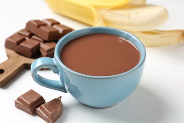 Tasty chocolate milk in cup, pieces and banana on white wooden table, closeup
