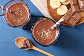 Tasty chocolate milk in glasses, pieces and banana on blue wooden table, flat lay
