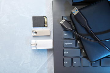 Flash drives, SD memory card, external hard disk and laptop on gray textured table, flat lay