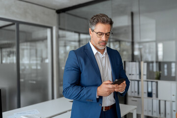 Focused Indian mature businessman in suit and eyeglasses holding smartphone standing in office,...