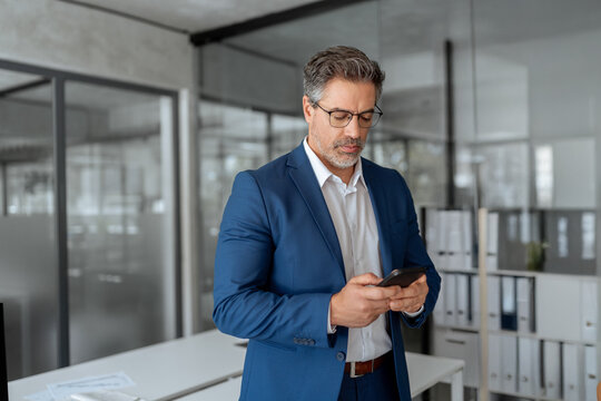 Focused Indian mature businessman in suit and eyeglasses holding smartphone standing in office, working on device. Middle eastern arab or latin senior business manager man ceo using cell phone mobile