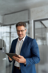 Fototapeta premium Focused senior mature Indian or Latin entrepreneur businessman holding digital pc tablet standing in office. Smiling hispanic man in suit working using touchpad computer for business work. Vertical 