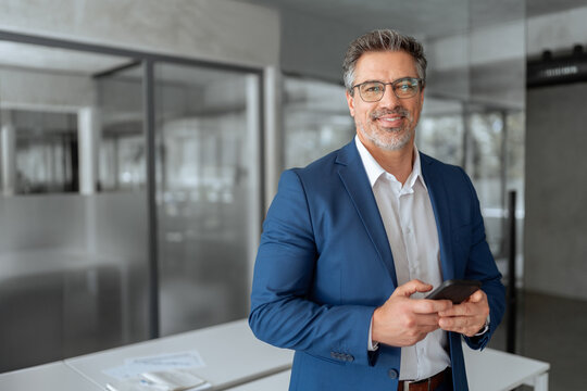 Smiling Indian mature businessman in suit holding smartphone standing in office, working on device. Executive professional ceo senior business manager man using cell phone mobile, looking at camera