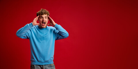 Young man holding hands on head and open moth in shock against red studio background with copy space. Concept of wow, sale, emotions, fashion, style, surprise. Ad