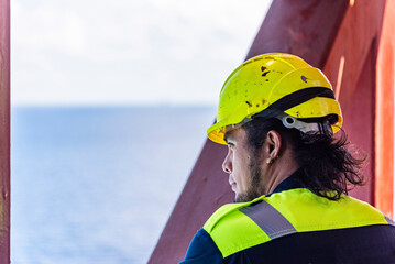Portrait of a young Filipino officer in protective clothing and a yellow helmet, standing aboard a container ship - his eyes focused ahead, reflecting both duty and quiet determination.
