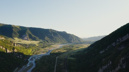 Panoramic shooting from a quadcopter. View of a mountain valley bathed in sunlight. Green hills overgrown with forest at foot of the rocky mountains. Mountain river in the lowland between mountains
