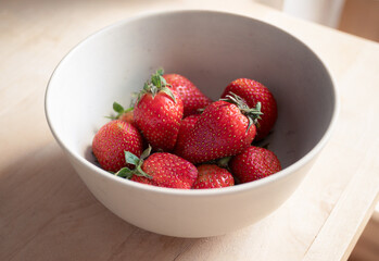 Fresh strawberries in a bowl resting on a wooden table, bathed in warm natural light. The vibrant red fruit stands out against the soft tones of the wood, creating a cozy and appetizing composition.