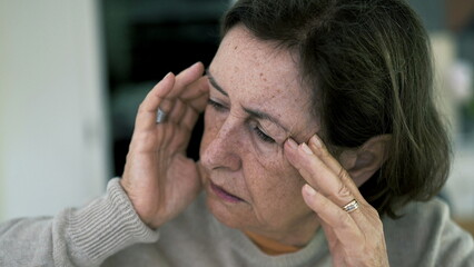 Senior woman in visible distress, holding head with eyes closed, fighting severe headache. Facial...
