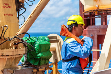 Merchant ship officer in blue overalls and yellow helmet, wearing lifejacket,  explains lifeboat...