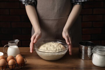 Woman with bowl of raw dough and ingredients at wooden table, closeup