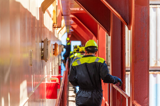 Deck officer seen from behind walking between containers and superstructure during an abandon-ship drill, dressed in blue coveralls and yellow helmet - real action on a working merchant ship.