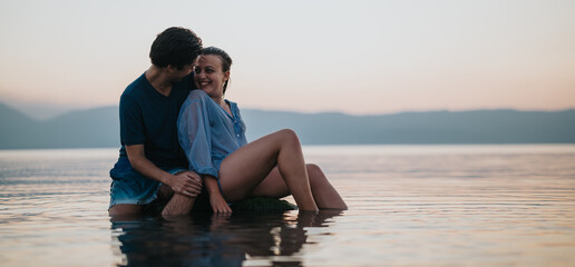 A loving couple sits closely together in calm water during a picturesque sunset. The scene exudes...